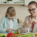 Helpful daughter is making vegetable salad chopping cucumber while young loving mother is touching her hair expressing love at home in kitchen.